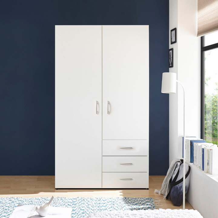 A white 2-door wardrobe with drawers and shelves, placed against a blue wall in a room with a window and a lamp.