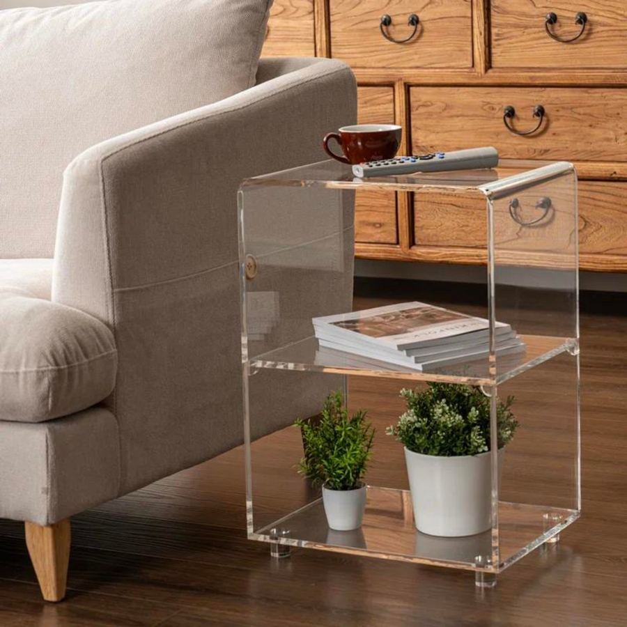 A clear acrylic three-tier end table with round raised feet, placed on a wooden floor next to a beige sofa, with items on the table including a plant in a white pot and a remote control.