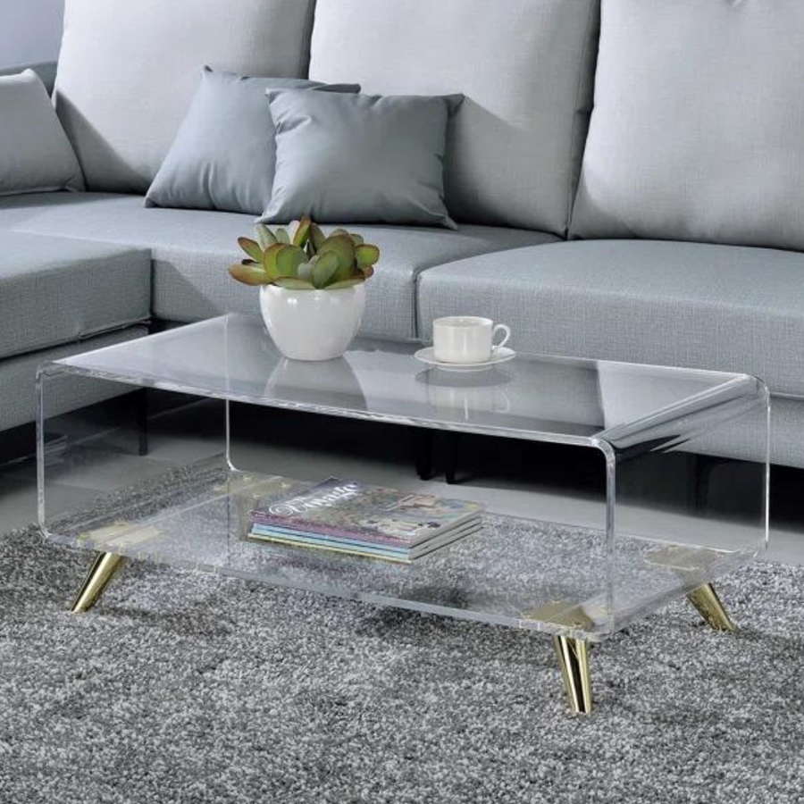 Clear acrylic coffee table with a waterfall-edge design and angled gold metal legs, placed on a grey area rug in a living room setting with a white vase on top, a cup, and some magazines.