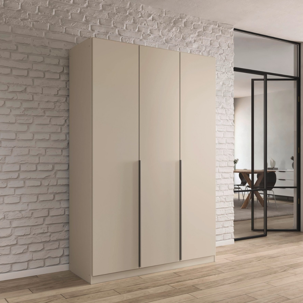 A beige 3-door wardrobe with a minimalist design, placed against a brick wall in a modern interior setting. The wardrobe has a glossy finish and handle-less doors.