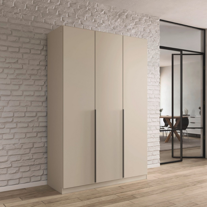 A beige 3-door wardrobe with a minimalist design, placed against a brick wall in a modern interior setting. The wardrobe has a glossy finish and handle-less doors.