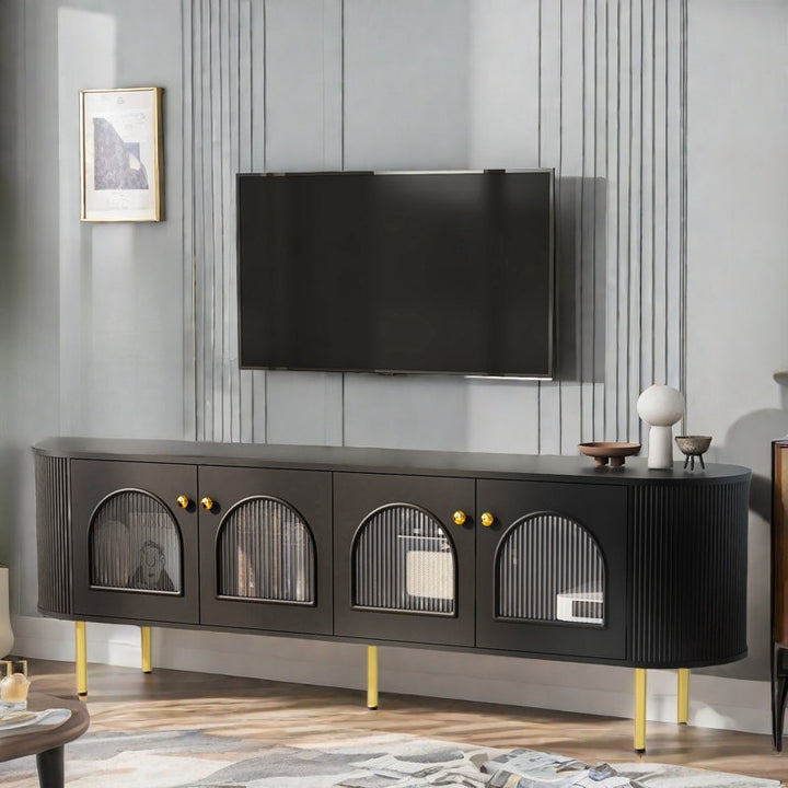 A black Mint TV stand with curved edges and fluted glass shutters, featuring golden handles and knobs, and standing on gold legs, with a flat-screen TV on top.