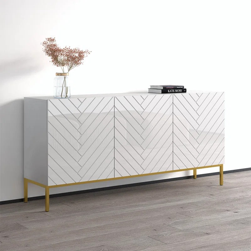 A modern white sideboard with geometric patterned panels and gold-colored legs, featuring open shelves and closed shutters. There is a vase with flowers on top and a book on the sideboard.
