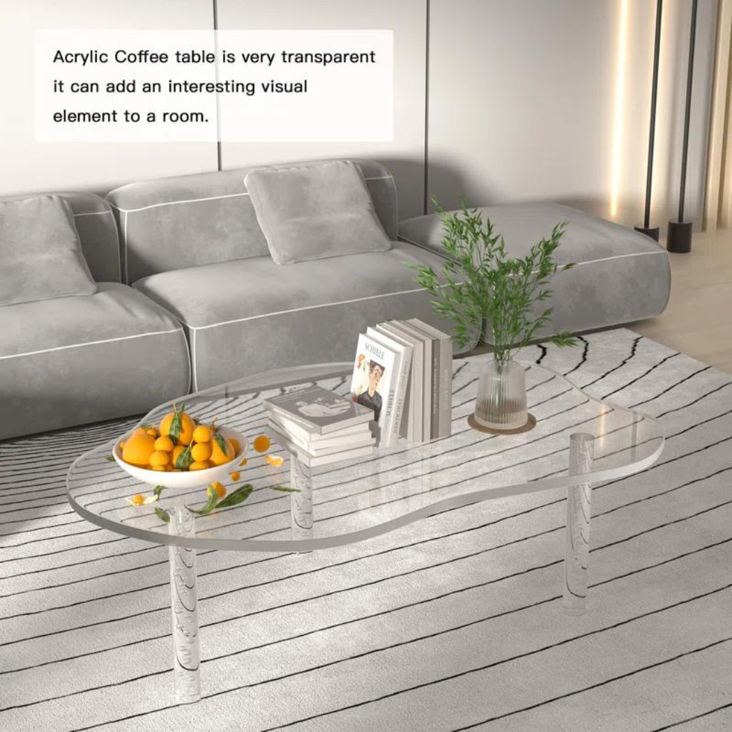A clear acrylic coffee table with a unique cloud-shaped design, featuring bubble-adorned legs and placed on a striped rug next to a sofa in a modern living room setting, with decorative items and books on top.