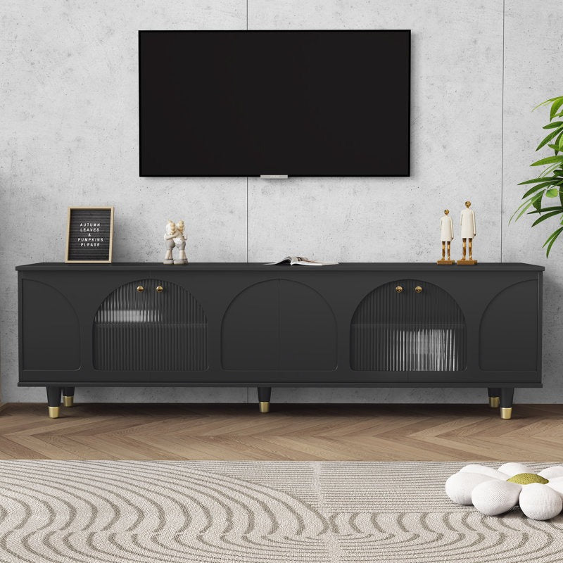 A large black TV stand with fluted glass doors and golden accents, mounted on white and gold legs. It features a high-gloss PU finish and is positioned in a room with a flat-screen TV on top and decorative items and plants around.