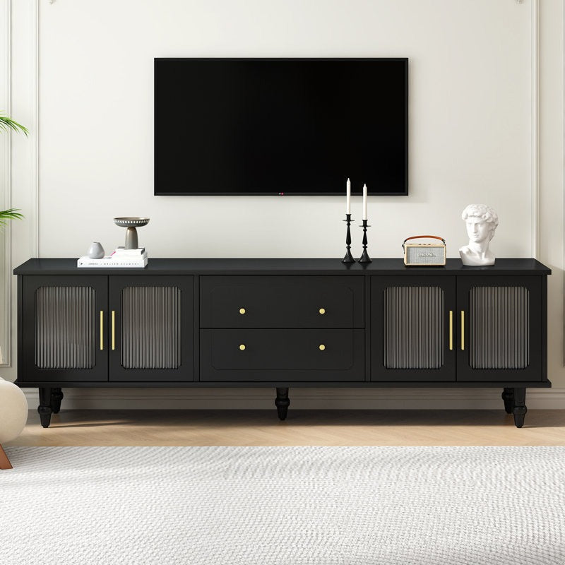 A black TV stand with fluted glass doors, golden handles, and white legs against a white wall in a room with a flat-screen TV on top and decorative items placed on it.