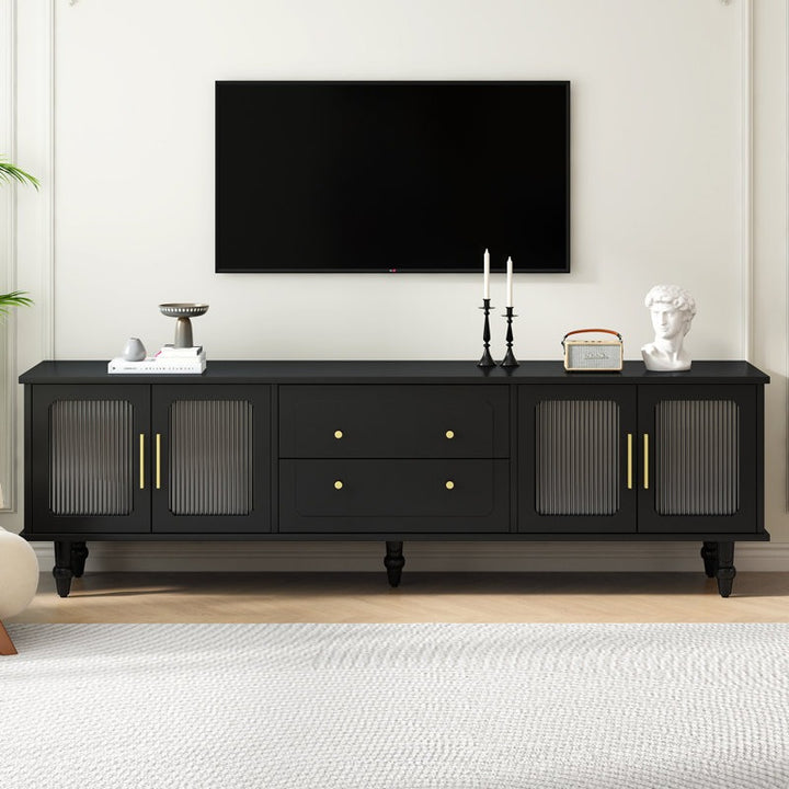 A black TV stand with fluted glass doors, golden handles, and white legs against a white wall in a room with a flat-screen TV on top and decorative items placed on it.