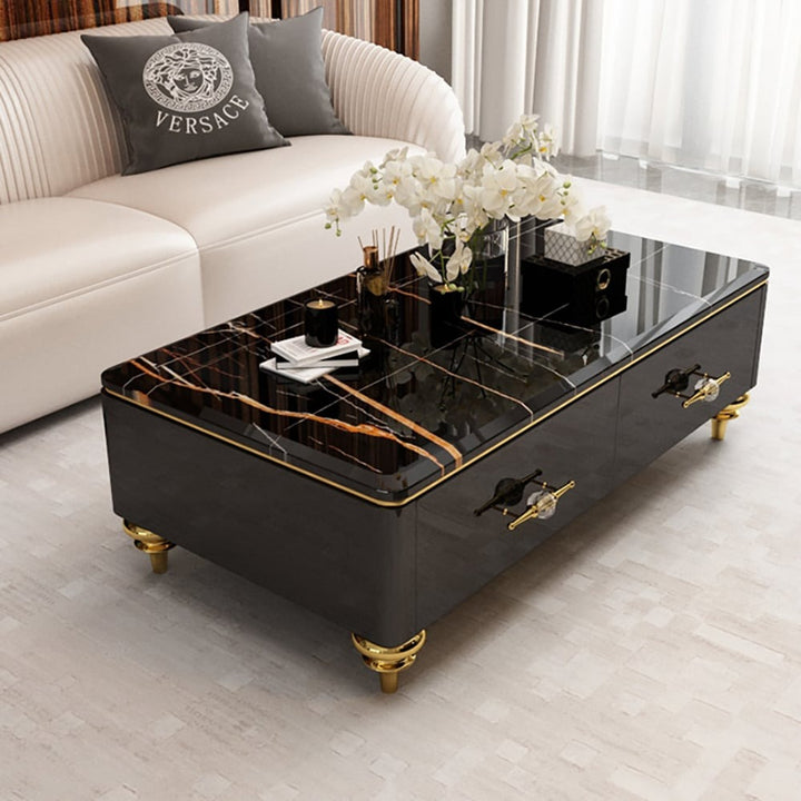 A modern neoclassic black coffee table with a luxurious marble top, gold-colored metal legs, and four drawers, placed in a living room setting.