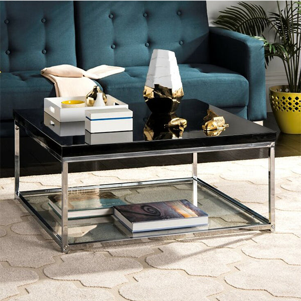 A black Marcale coffee table with a clear glass shelf, placed on a rug in a living room setting. The table has a stainless steel frame and is positioned against a blue upholstered sofa with decorative items on top.