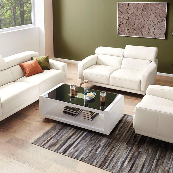A modern white coffee table with acrylic legs, placed on a grey area rug in a living room setting with two couches, a glass top, and various decorative items on top.