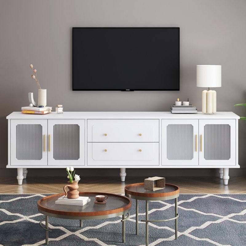 A white TV stand with fluted glass doors and golden handles, featuring a high-gloss PU-painted finish. It is placed in a room with a flat-screen TV on top, flanked by two round tables with a lamp and decorative items.