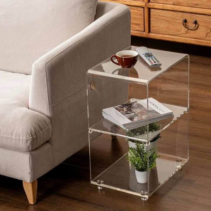 A clear acrylic three-tier end table with round raised feet, placed on a wooden floor next to a beige sofa, with items on the table including a plant in a white pot and a remote control.