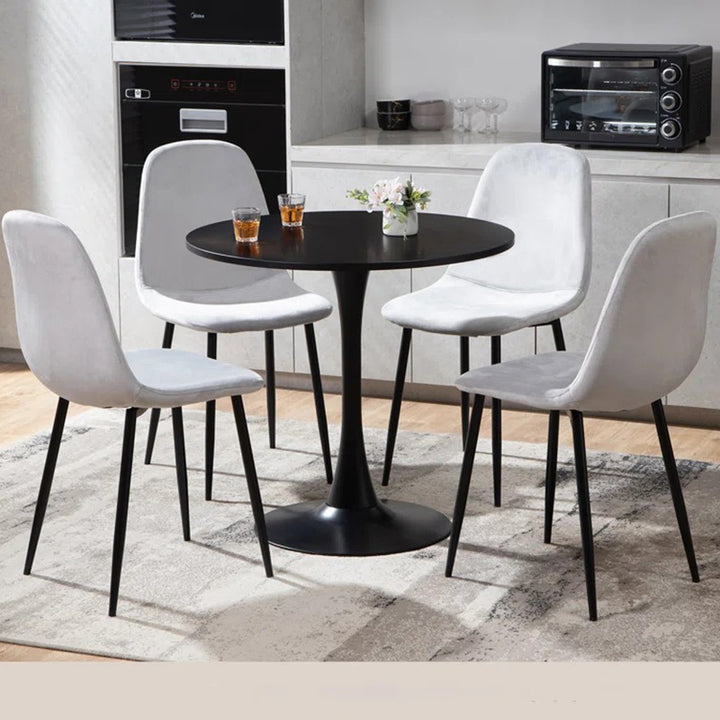 A modern dining set with a round table and four chairs, featuring white upholstered fabric seats and black metal legs, placed on a rug in a room with kitchen appliances in the background.