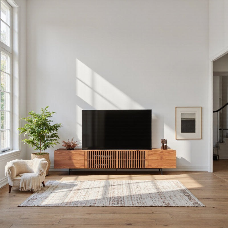 A large FILA TV stand made of rubber wood with metal legs, featuring four cabinets and visible hidden compartments, in a bright living room setting with a flat-screen TV on top, alongside a armchair and a side table with a lamp.