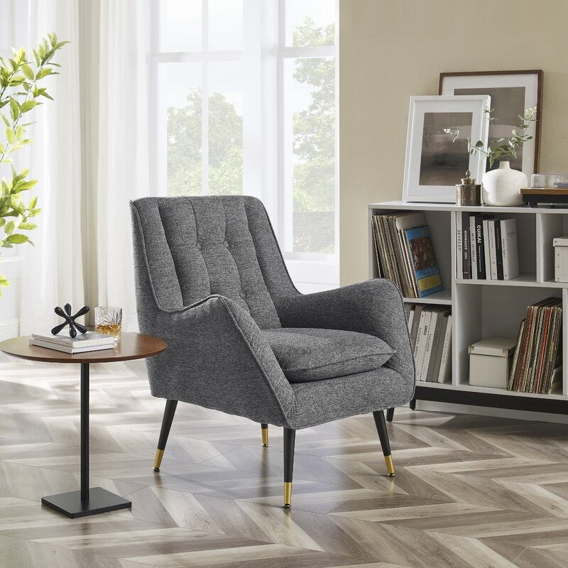 A gray upholstered Bruy Lounge Chair with a dramatic wingback silhouette, flared arms, and button tufting, positioned in a room with a herringbone wood floor and bookshelves in the background.