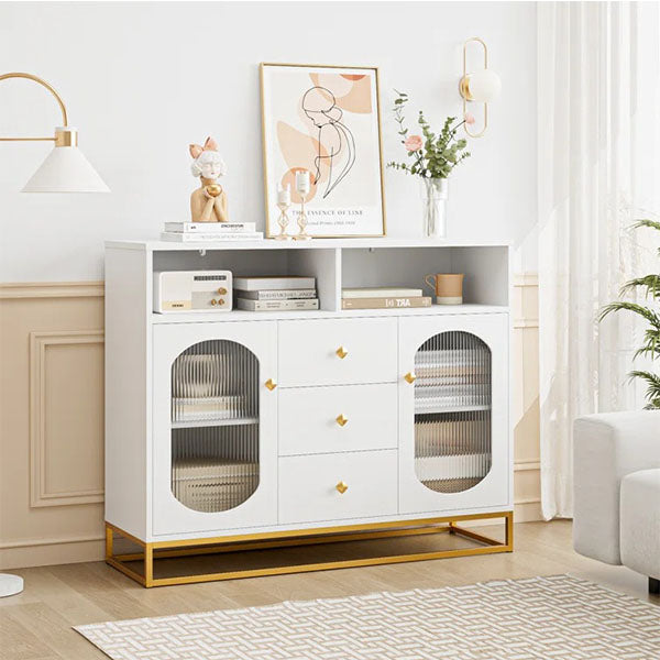 A white sideboard and cabinet with gold accents, featuring glass doors, open shelves, and drawers, are placed in a room with decorations and a plant.