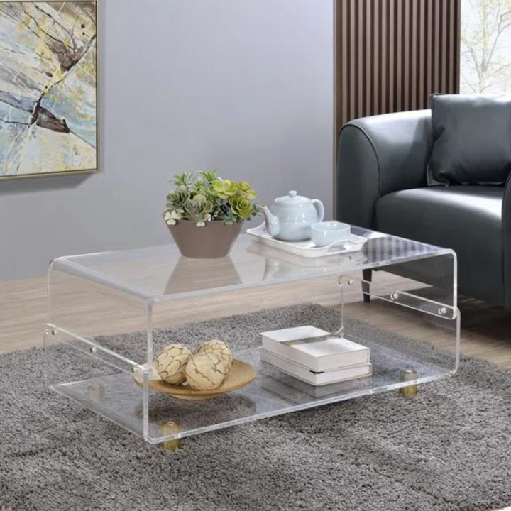 A clear acrylic coffee table with a waterfall edge design, featuring two-tier storage and gold metal legs, placed on a grey area rug in a living room setting with a couch in the background and decorative items on top.