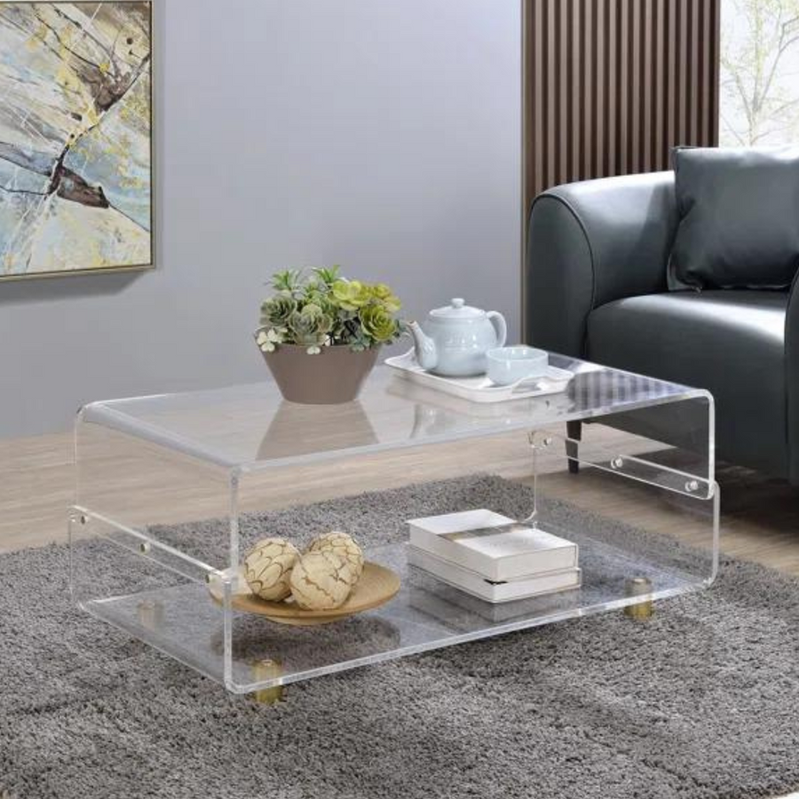 A clear acrylic coffee table with a waterfall edge design, featuring two-tier storage and gold metal legs, placed on a grey area rug in a living room setting with a couch in the background and decorative items on top.