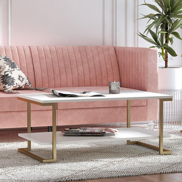 A white rectangular coffee table with gold metal legs, placed on a rug in a room with a pink velvet sofa and a plant in a pot.