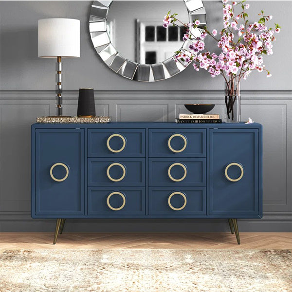 A navy blue sideboard with gold-colored ring-shaped handles and metal feet, featuring multiple drawers and cabinets, placed in a room with a mirror, lamp, and decorative items on top.