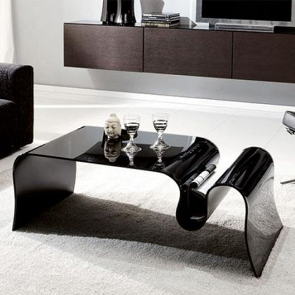 A black, acrylic coffee table with a smooth finish, placed on a white carpet in a living area setting, accompanied by two glasses and a book.