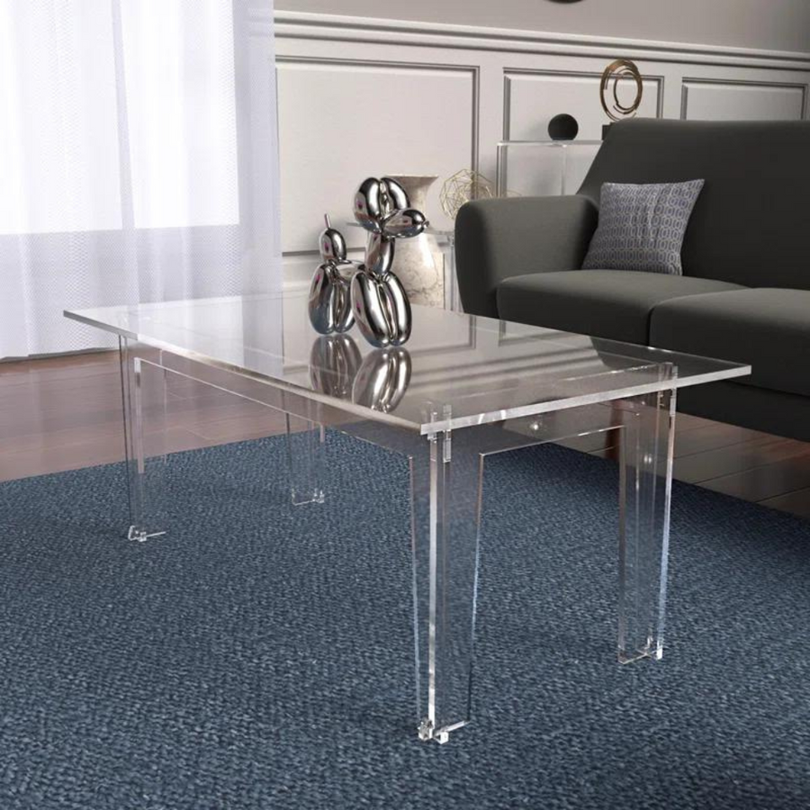 A clear acrylic coffee table with a waterfall-edge design placed on a blue area rug in a living room setting, with a couch and decorative items on top of the table.
