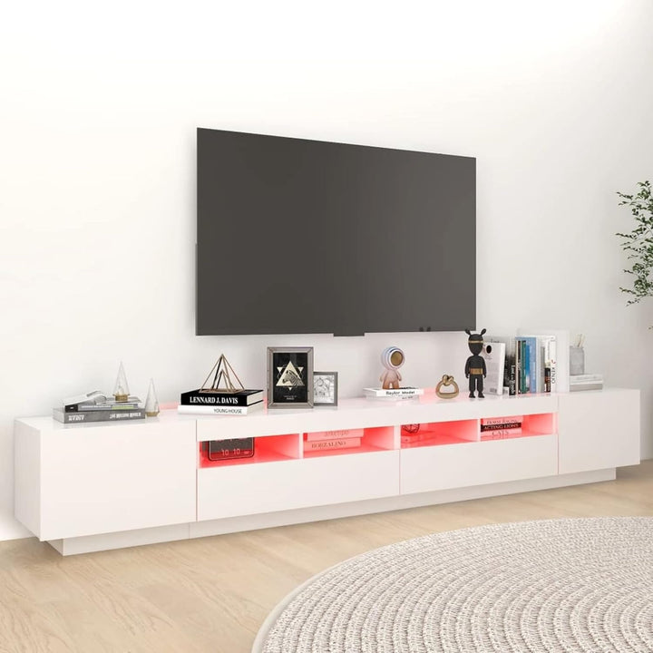 A modern white TV stand with ambient LED lighting, featuring shutters and raised metallic legs, with a flat-screen TV and various decorative items on top.
