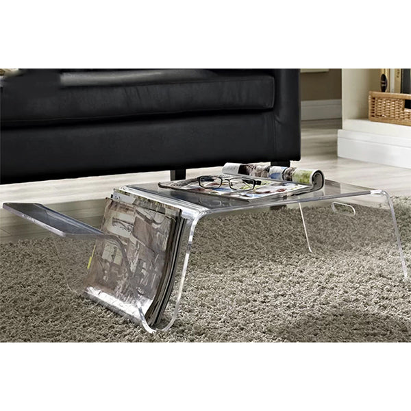 A clear acrylic coffee table with a magazine on top, positioned on a carpeted floor next to a black sofa with a wooden side table.