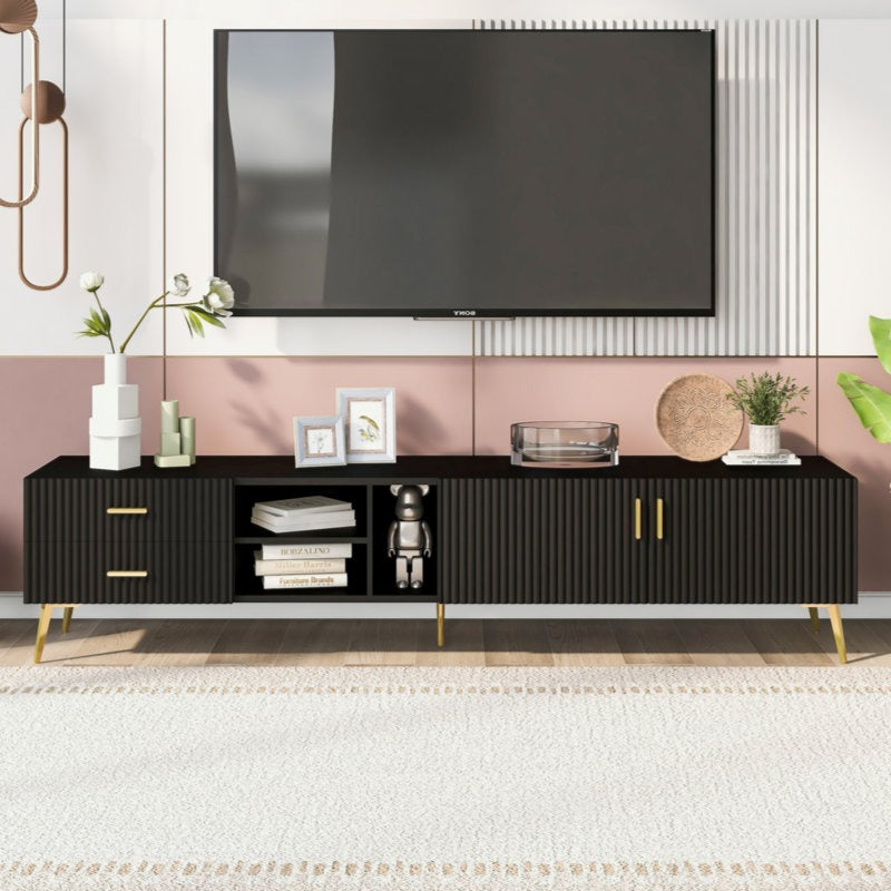 A modern black TV stand with hidden compartments, open shelves, and gold hardware. It is positioned in a room with a large screen TV on top, decorative items, and plants.