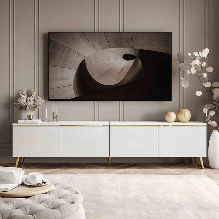 A white high-gloss TV stand with golden accents and metal legs, featuring four shutter compartments, placed in a room with a wall-mounted TV on top.