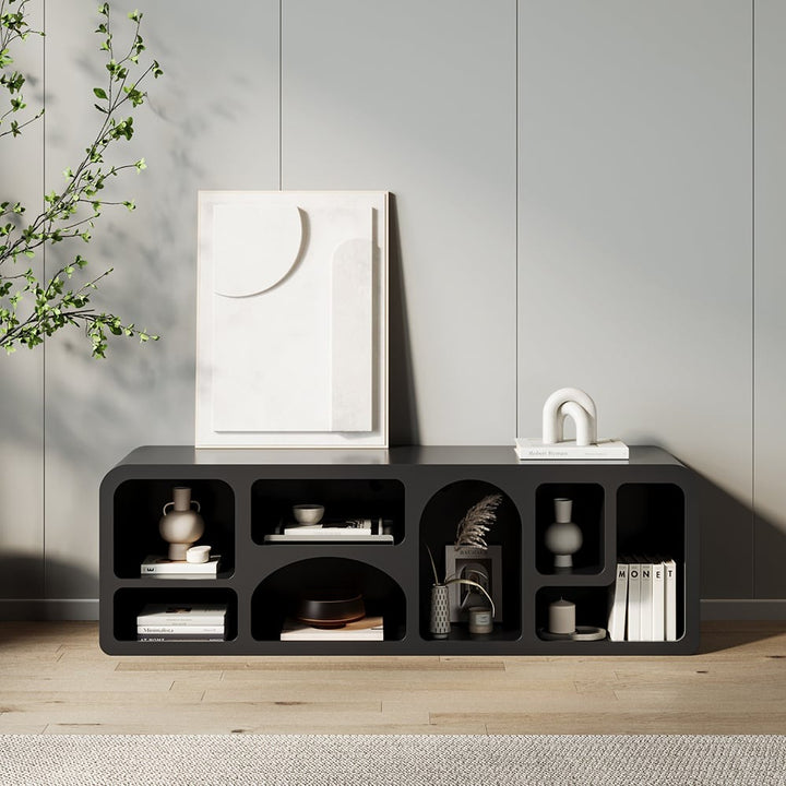 A modern black TV shelf unit with various items placed on it, set against a light-colored wall with a plant to the side.