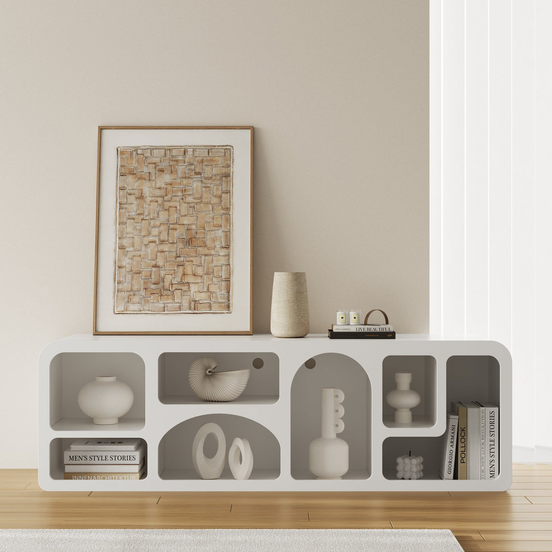 A modern white TV shelf unit with various decorative items displayed on it, set against a neutral wall in a bright room.