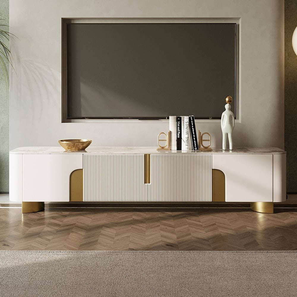Modern white TV stand with symmetrical cabinet doors and stone top, featuring gold accents and rounded corners, set in a room with a large screen TV on top.