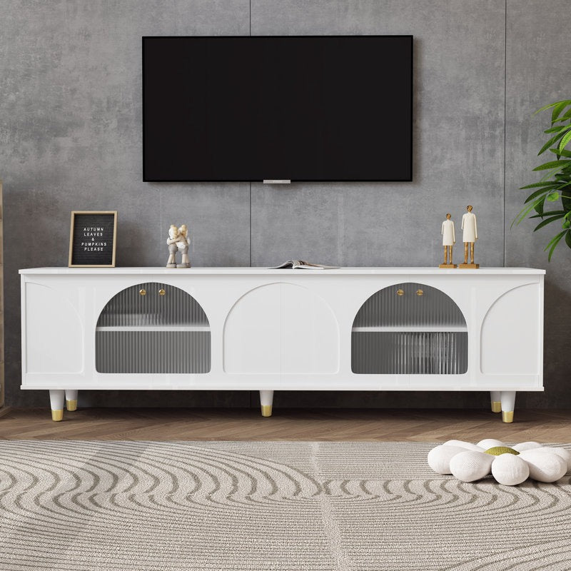 A white TV stand with fluted glass doors and golden accents, mounted on white-gold legs. A television and decorative items are placed on top, set against a minimalist interior background.