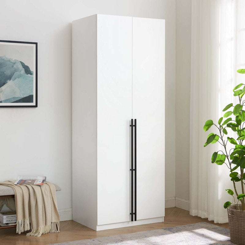 A white 2-door wardrobe with minimal design, featuring black vertical handles and standing in a room with a plant and a framed picture on the wall.