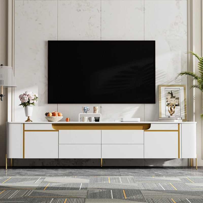 Modern white TV stand with gold accents, featuring multiple storage drawers and shutters, supporting a flat-screen TV on top, placed in a room with decorative items and a window in the background.