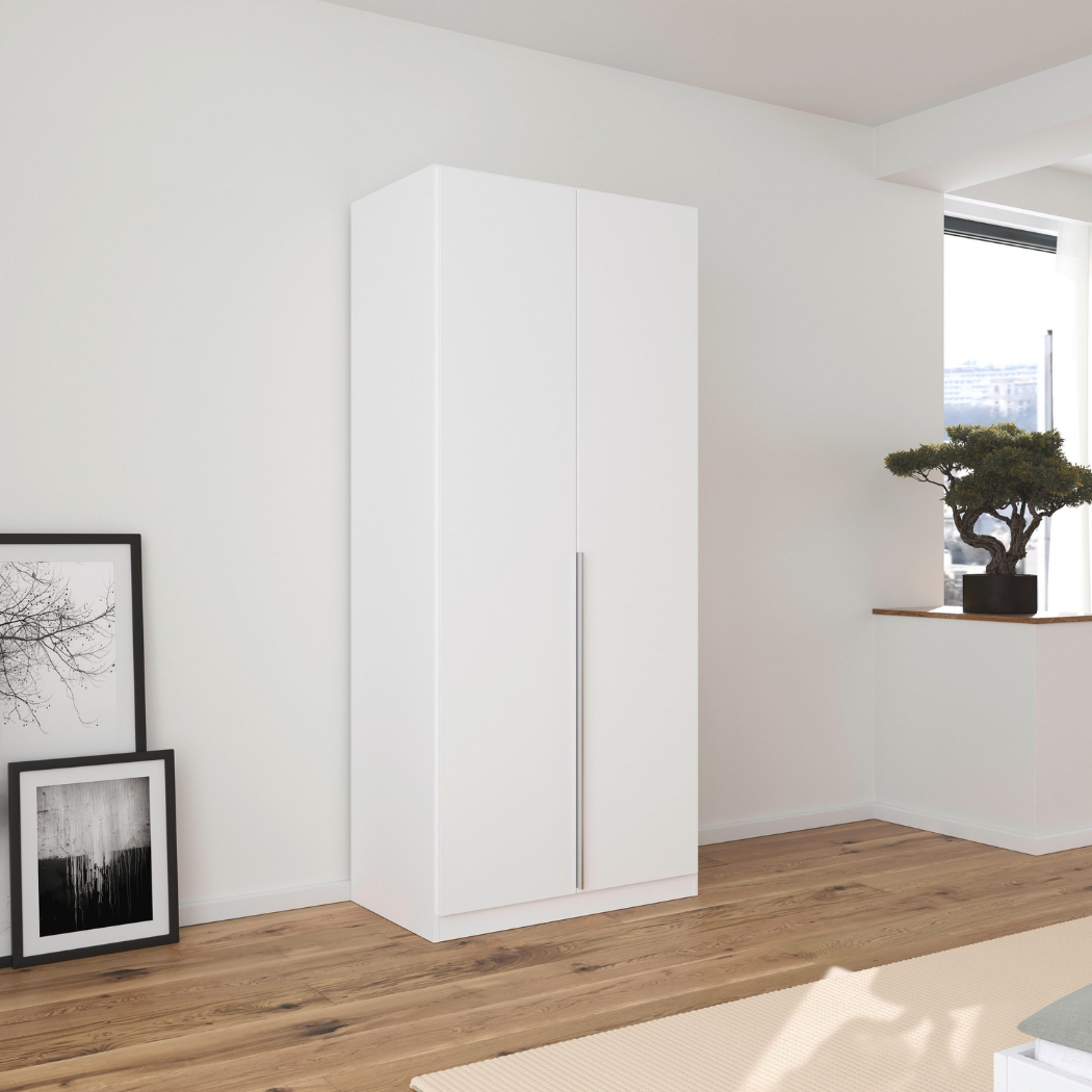 A matte white 2-door wardrobe with integrated handles, placed against a minimalist background with a framed picture and a small plant to the side.