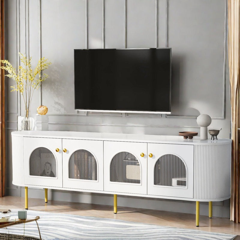 A white Mint TV stand with curved edges and golden handles, featuring fluted glass shutters, positioned in a room with a mounted television on top.