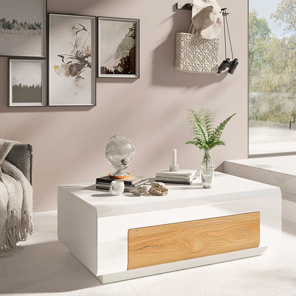 A modern white coffee table with a wooden drawer, placed in a bright living area with decorative items and a mirror on top.