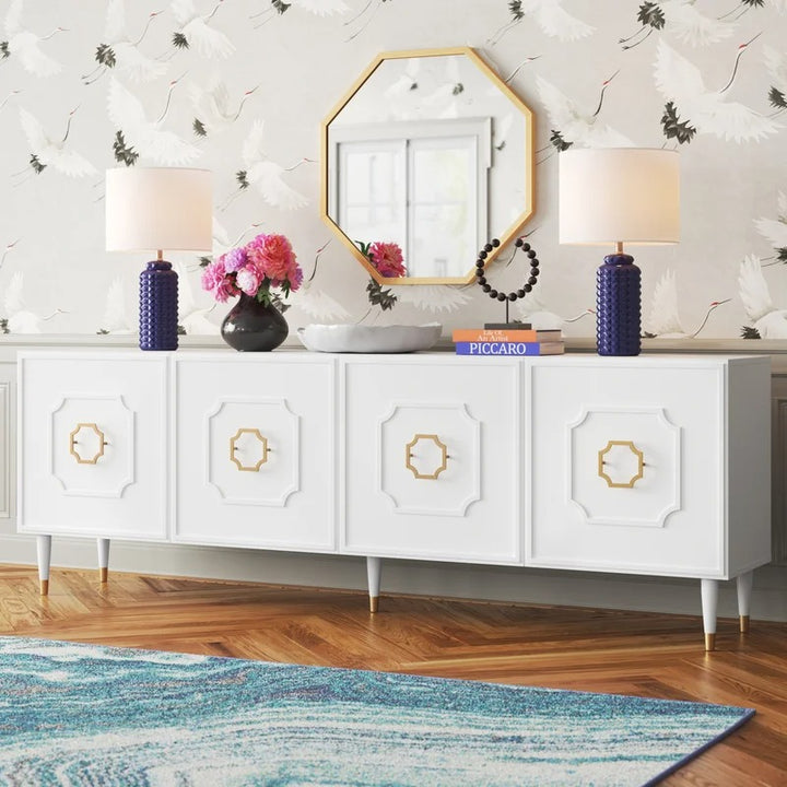 A white sideboard with gold handles and a mirror above it, set against a wall with a floral pattern and a blue area rug. The white sideboard has a rectangular top and four doors, with decorative items and a lamp on top.