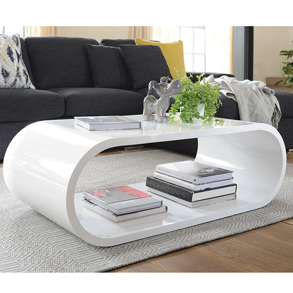 A white, modern elliptical coffee table made of wood, placed on a rug in a living room setting, with books and a plant on top.