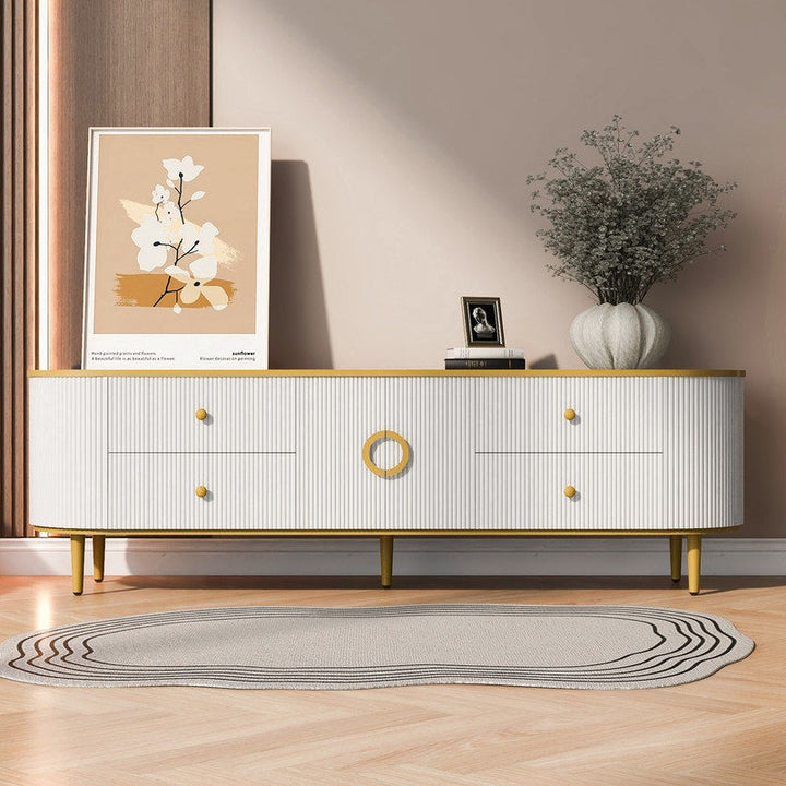 Modern white TV stand with golden details and legs, featuring four drawers and two open shutters, placed in a room with a framed picture, a plant, and a framed photo.