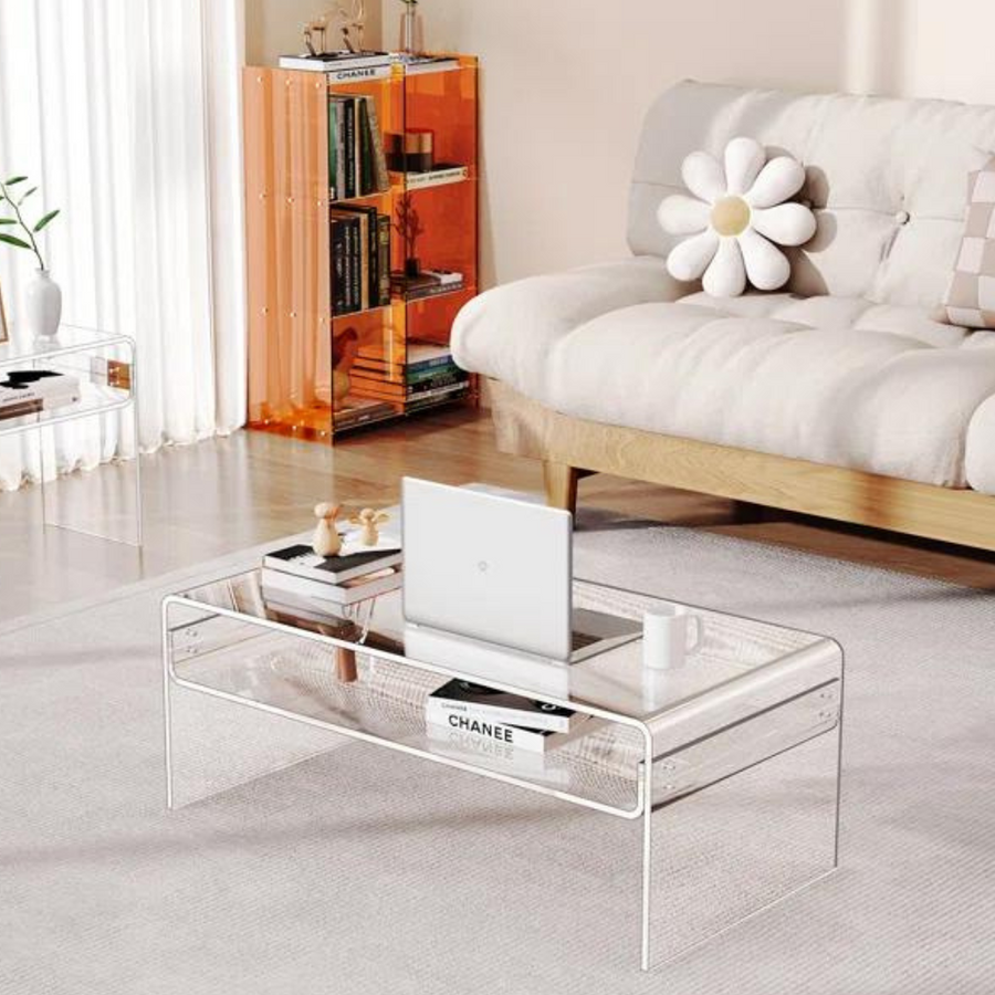 A clear acrylic coffee table with a waterfall-edge design placed on a carpeted floor next to a beige sofa, with a laptop and a flower vase on top of the table.